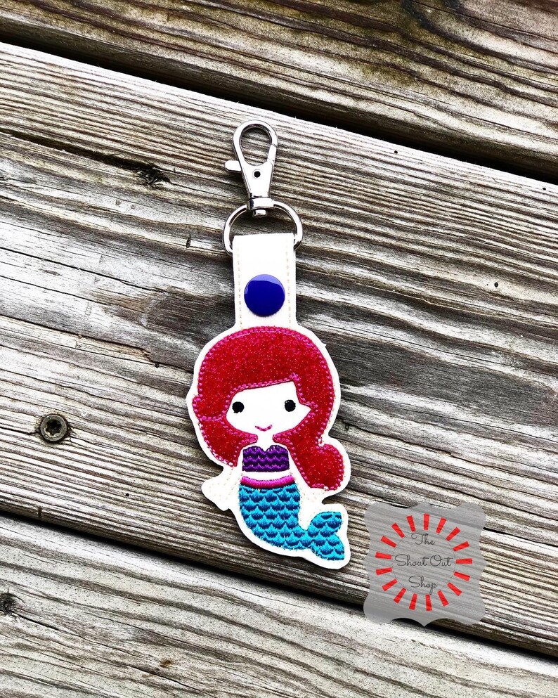 May include: A white keychain with a red-haired mermaid with a blue tail and a purple top. The keychain has a silver clip and a blue button. The text "The Short Cut Shop" is visible in the bottom right corner of the image.