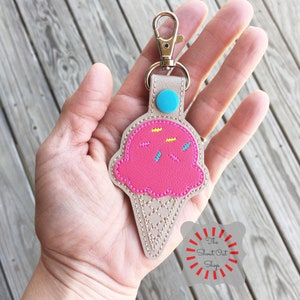 Puede incluir: Un llavero de cono de helado rosa con chispas amarillas y azules. El llavero est&aacute; hecho de fieltro y tiene un llavero de metal plateado. El llavero est&aacute; sostenido en una mano. El texto "The Shout Out Shop" es visible en la esquina inferior derecha de la imagen.