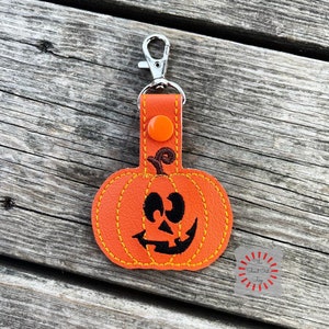 May include: Orange pumpkin-shaped keychain with a stitched black jack-o'-lantern face. The keychain has a silver metal loop and a red button.