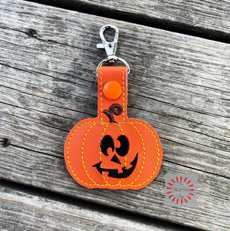May include: Orange pumpkin-shaped keychain with a stitched black jack-o'-lantern face. The keychain has a silver metal loop and a red button.