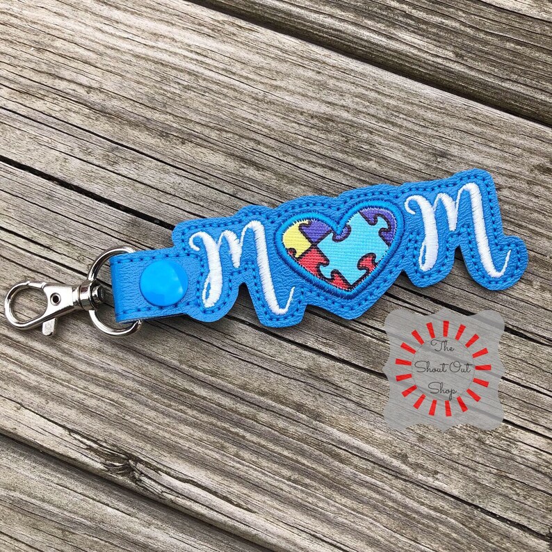 May include: Blue keychain with the word "MOM" in white cursive lettering. A heart with a blue puzzle piece design is in the center of the keychain.