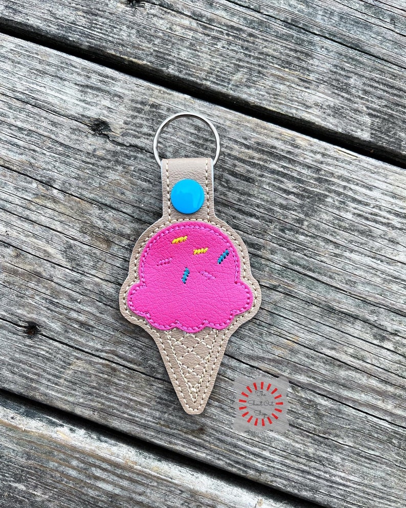 Puede incluir: Un llavero de cono de helado rosa con chispas amarillas y azules. El llavero est&aacute; hecho de cuero sint&eacute;tico y tiene un anillo de metal.