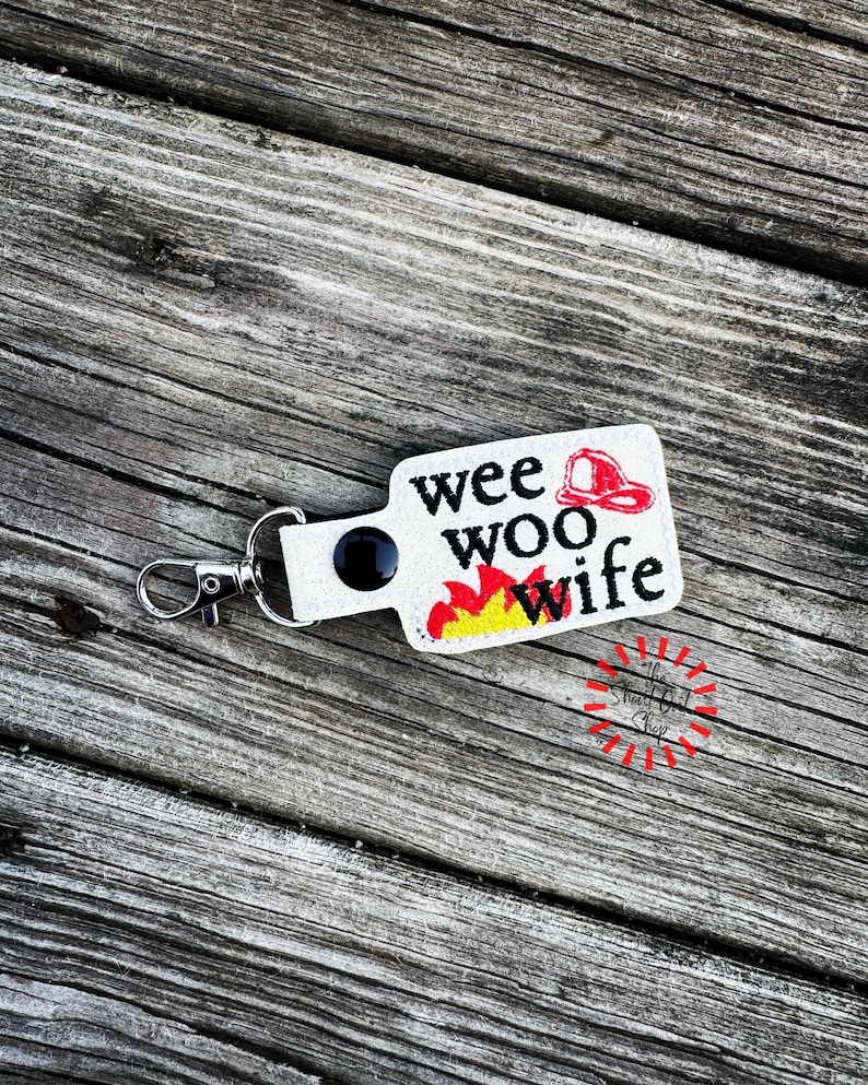 May include: A white rectangular keychain with the words "wee woo wife" in black text, a red firefighter's helmet, and flames in red and yellow. The keychain has a silver clasp and a black circular accent.