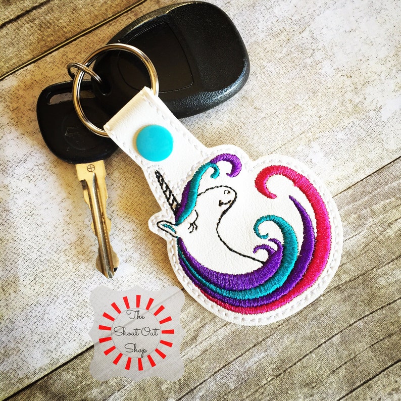 May include: White key chain with a colorful embroidered unicorn design. The unicorn has a purple, teal, and pink mane and tail. The key chain has a blue button and a silver key ring.