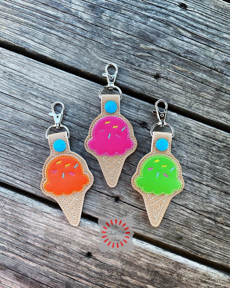Puede incluir: Tres llaveros con forma de cono de helado con helados de diferentes colores. El helado es rosa, naranja y verde. Cada llavero tiene un bot&oacute;n azul y un anillo de metal plateado.