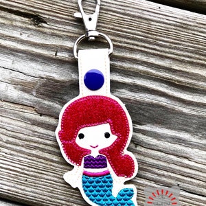 May include: A red-haired mermaid keychain with a blue tail and purple top. The keychain has a white background and a blue button.