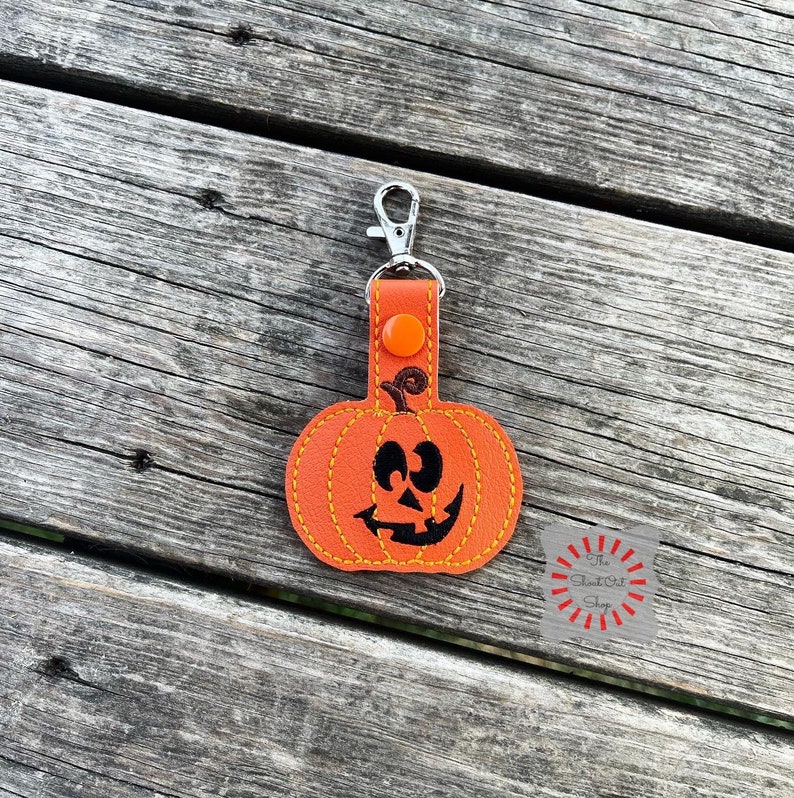 May include: Orange pumpkin-shaped keychain with a black stitched-on face. The keychain has a silver metal clasp and a small orange button.