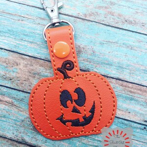 May include: Orange leather pumpkin keychain with a black stitched jack-o'-lantern face. The keychain has a silver metal clasp.