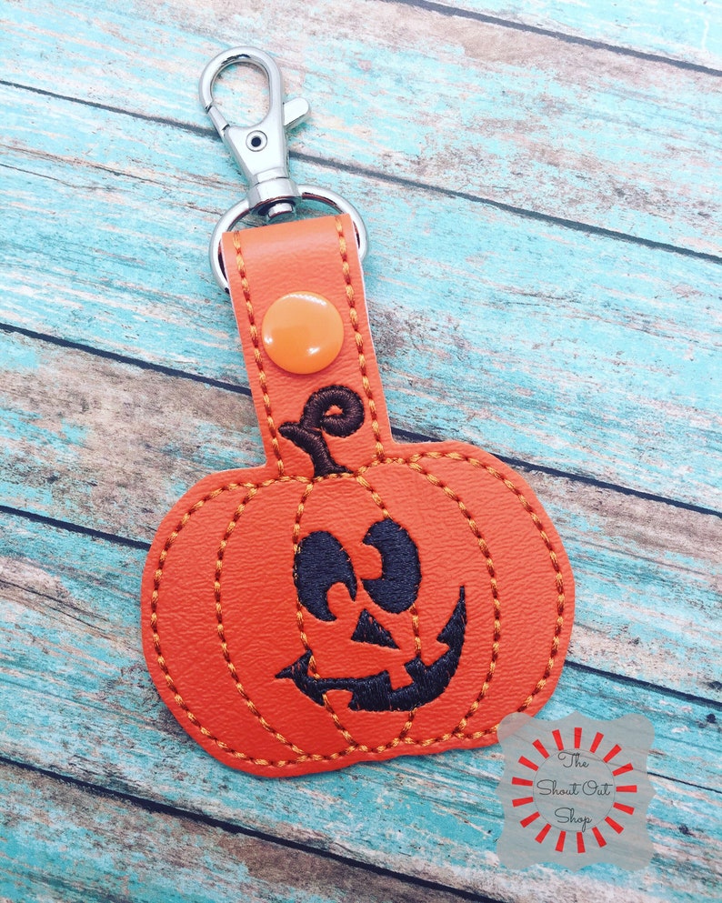 May include: Orange leather pumpkin keychain with a black stitched jack-o'-lantern face. The keychain has a silver metal clasp.