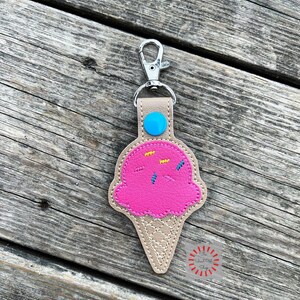 Puede incluir: Un llavero de cono de helado rosa con chispas y un bot&oacute;n azul en la parte superior. El llavero est&aacute; hecho de cuero y tiene un cierre de metal plateado.