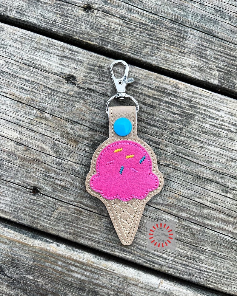 Puede incluir: Un llavero de cono de helado rosa con chispas y un bot&oacute;n azul en la parte superior. El llavero est&aacute; hecho de cuero y tiene un cierre de metal plateado.
