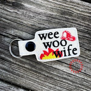 May include: A white glitter keychain with the words "wee woo wife" in black, a red firefighter's helmet, and flames in yellow and red. The keychain has a black button and a silver key ring.