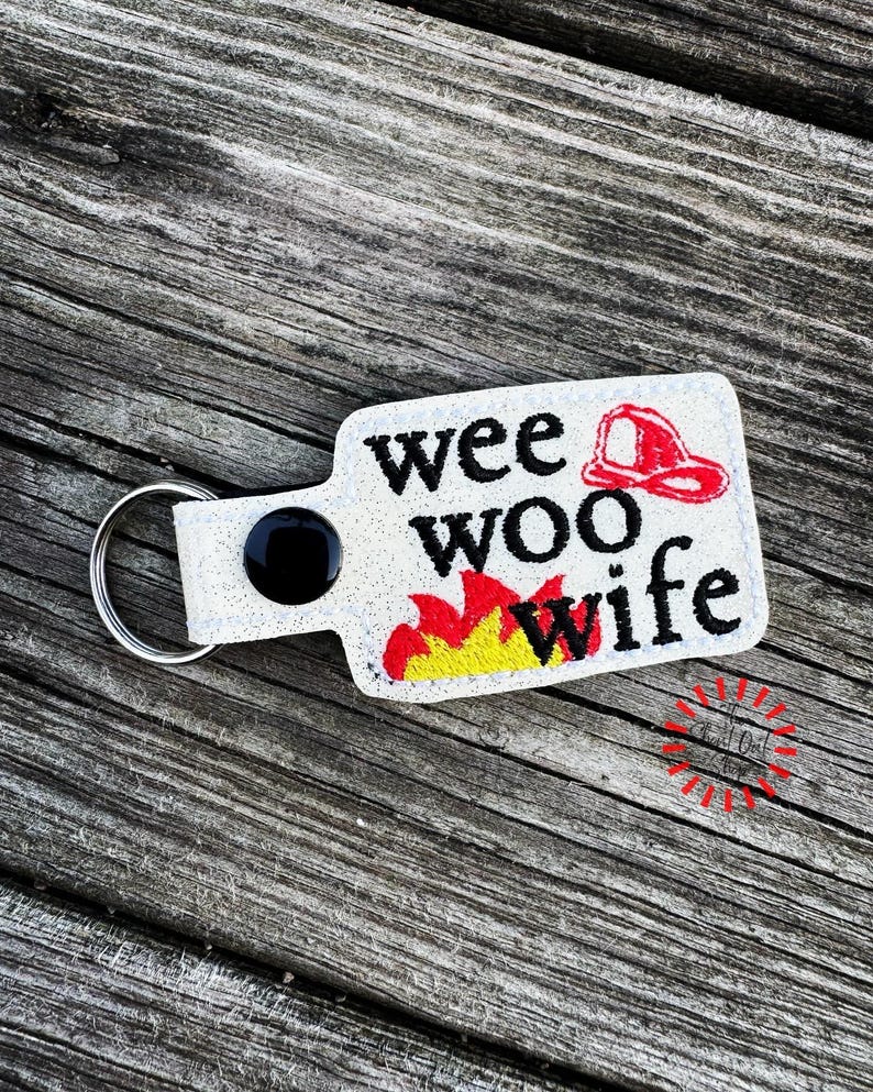 May include: A white glitter keychain with the words "wee woo wife" in black, a red firefighter's helmet, and flames in yellow and red. The keychain has a black button and a silver key ring.
