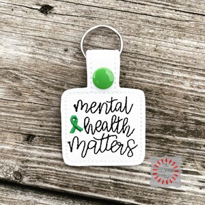 May include: White keychain with a green button and a green ribbon. The keychain has the text "mental health matters" embroidered on it.