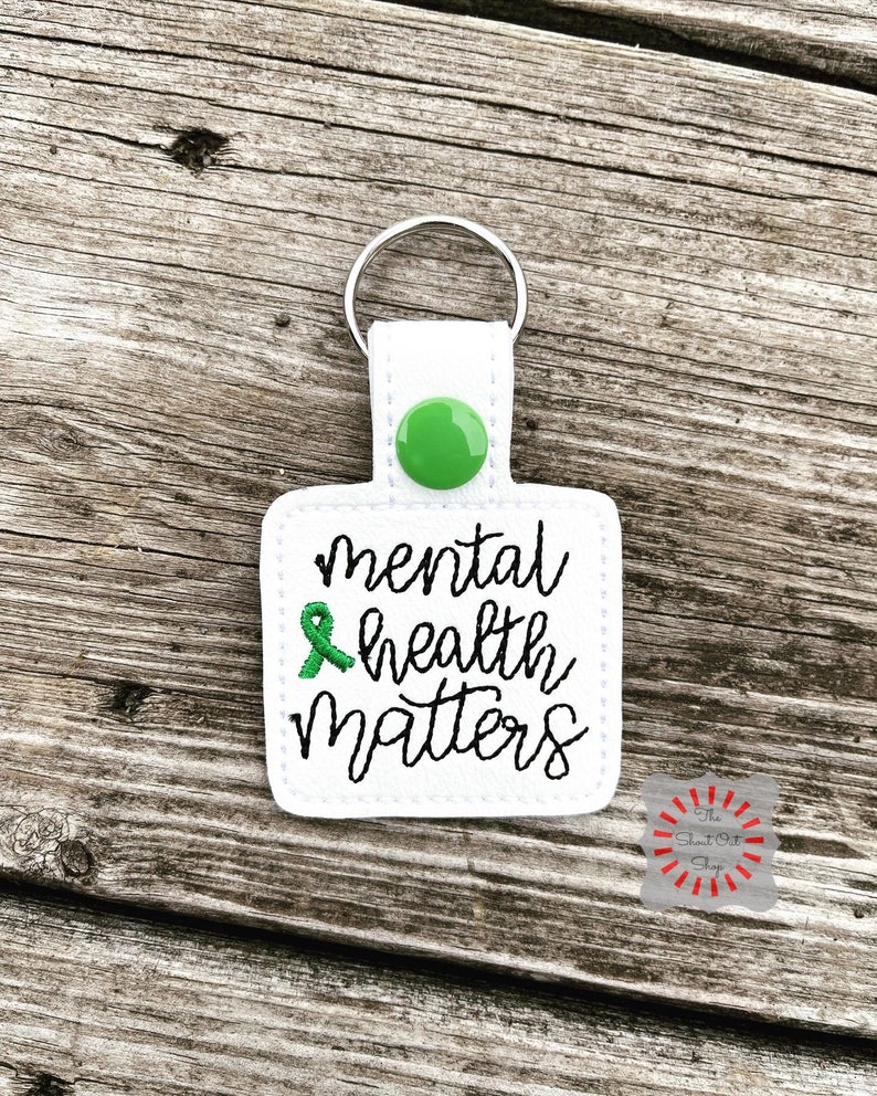 May include: White keychain with a green button and a green ribbon. The keychain has the text "mental health matters" embroidered on it.