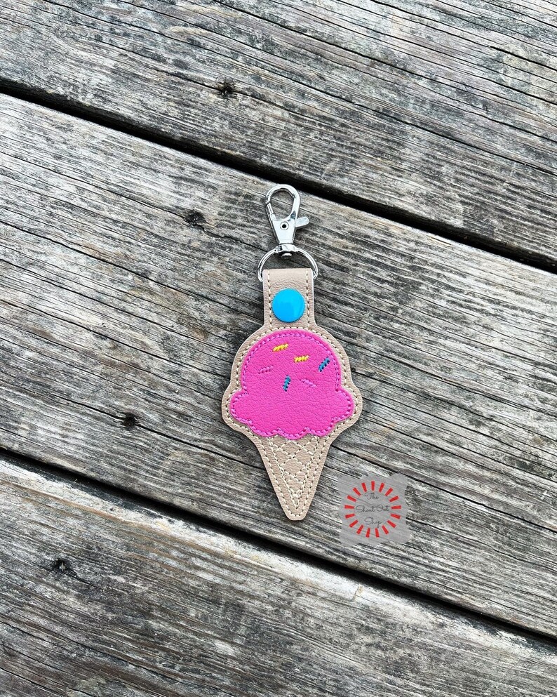 Puede incluir: Un llavero de cono de helado rosa con chispas azules y amarillas. El llavero est&aacute; hecho de fieltro y tiene un cierre de metal plateado.
