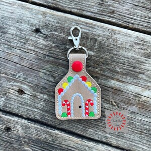 May include: A brown leather gingerbread house keychain with red, white, and green embroidery. The keychain has a silver metal clasp.