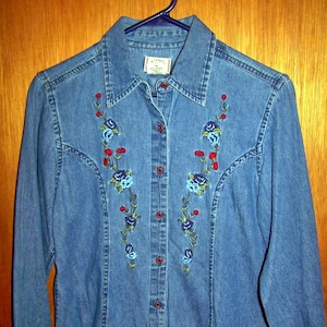 May include: A blue denim shirt with a floral embroidery design. The shirt has a button-up front and long sleeves.