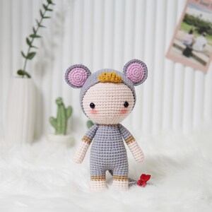 May include: A handmade crochet doll in the shape of a mouse. The doll is wearing a gray jumpsuit and a matching hood with pink ears. It has a small gold crown and a red tail. The doll is on a white surface.