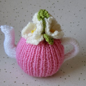 May include: A pink knitted teapot with white crocheted flowers and green leaves on top.