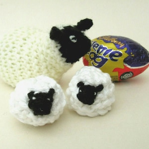 May include: Three white crocheted sheep with black faces. The largest sheep is standing next to a purple and gold foil-wrapped chocolate egg with the text "Cadbury Creme Egg".