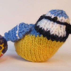May include: A knitted bluebird toy with a yellow belly, blue wings, and a white and black head. The bird has a black bead for an eye.
