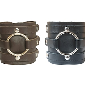 May include: Two leather bracelets with silver metal accents. One bracelet is brown and the other is black. Both bracelets have a large silver ring in the center.