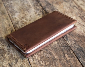 Puede incluir: Una funda de teléfono de cuero marrón con un borde cosido y las iniciales "ec" grabadas en la parte delantera. La funda es rectangular y parece que contiene un teléfono. La funda está sobre una superficie de madera.