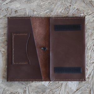May include: Open brown leather wallet with a card slot and a button closure. The wallet has a contrasting lighter brown interior and two black velcro strips for closure. The edges are stitched with a darker brown thread.