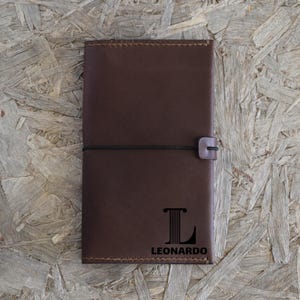 May include: A brown leather journal with a black elastic closure. The journal has the name "LEONARDO" and a stylized letter "L" printed on the front. The journal is closed and appears to be new.
