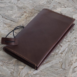 May include: A dark brown leather wallet with a stitched border and a black elastic closure. The wallet is rectangular and appears to be closed. The background is a light brown wood grain.