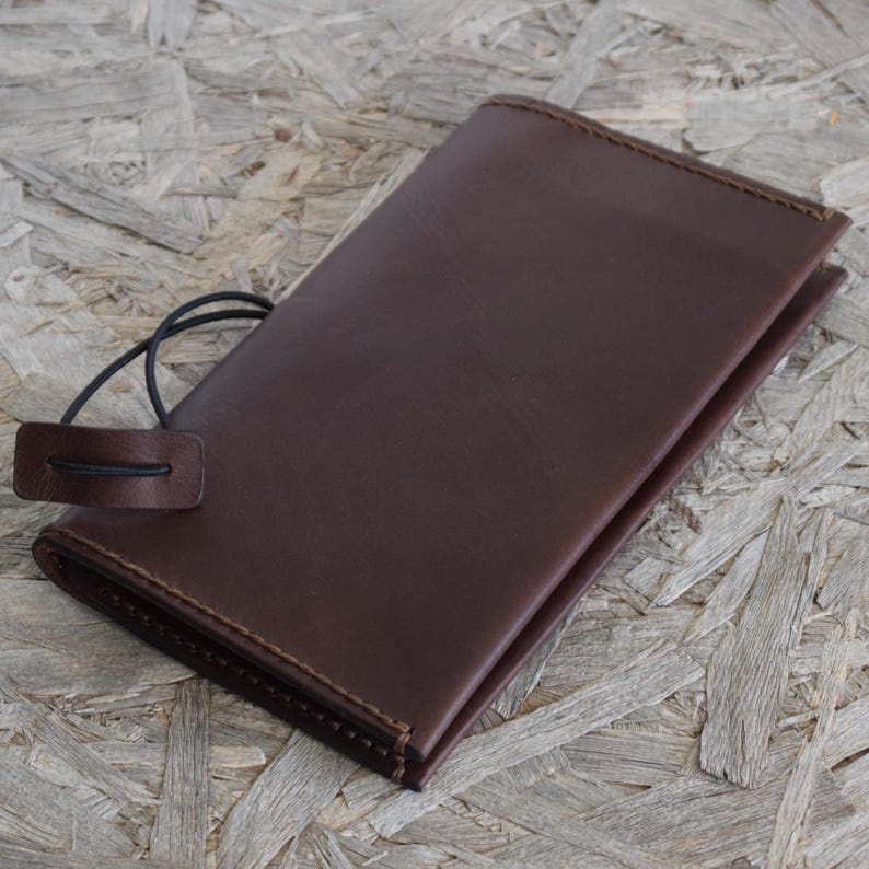May include: A dark brown leather wallet with a stitched border and a black elastic closure. The wallet is rectangular and appears to be closed. The background is a light brown wood grain.