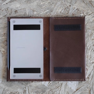 May include: A brown leather tablet case with a light-colored tablet inside. The case has a snap closure and two black velcro straps. A black stylus is visible on the left side. The tablet has the word "reMarkable" printed on it.