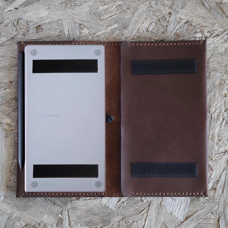 May include: A brown leather tablet case with a light-colored tablet inside. The case has a snap closure and two black velcro straps. A black stylus is visible on the left side. The tablet has the word "reMarkable" printed on it.