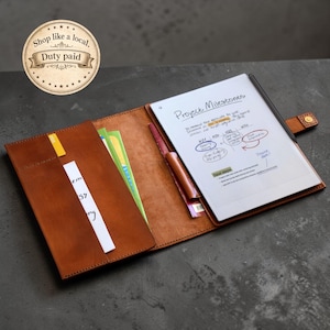 May include: A brown leather folio with a tablet and pen inside. The tablet displays handwritten notes with the title "Project Milestones." The folio has a snap closure and a pocket with a yellow tab. The text "Shop like a local. Duty paid" is in a circular emblem.