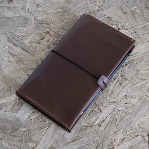 May include: A brown leather journal with a black elastic closure. The journal is rectangular and appears to be closed. The leather has a smooth texture. The journal is lying on a wooden surface.