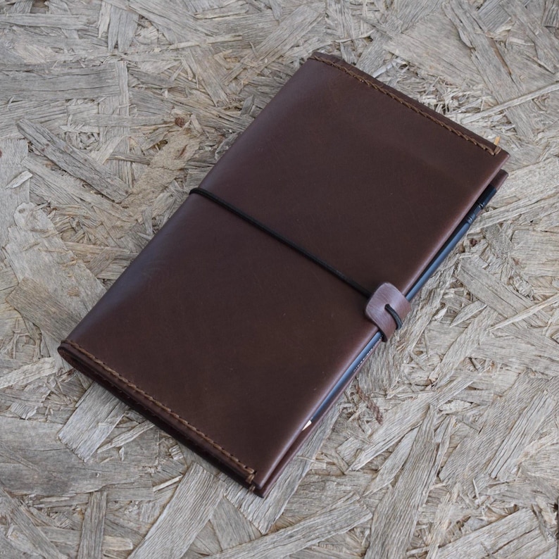 May include: A brown leather journal with a black elastic closure. The journal is rectangular and appears to be closed. The leather has a smooth texture. The journal is lying on a wooden surface.