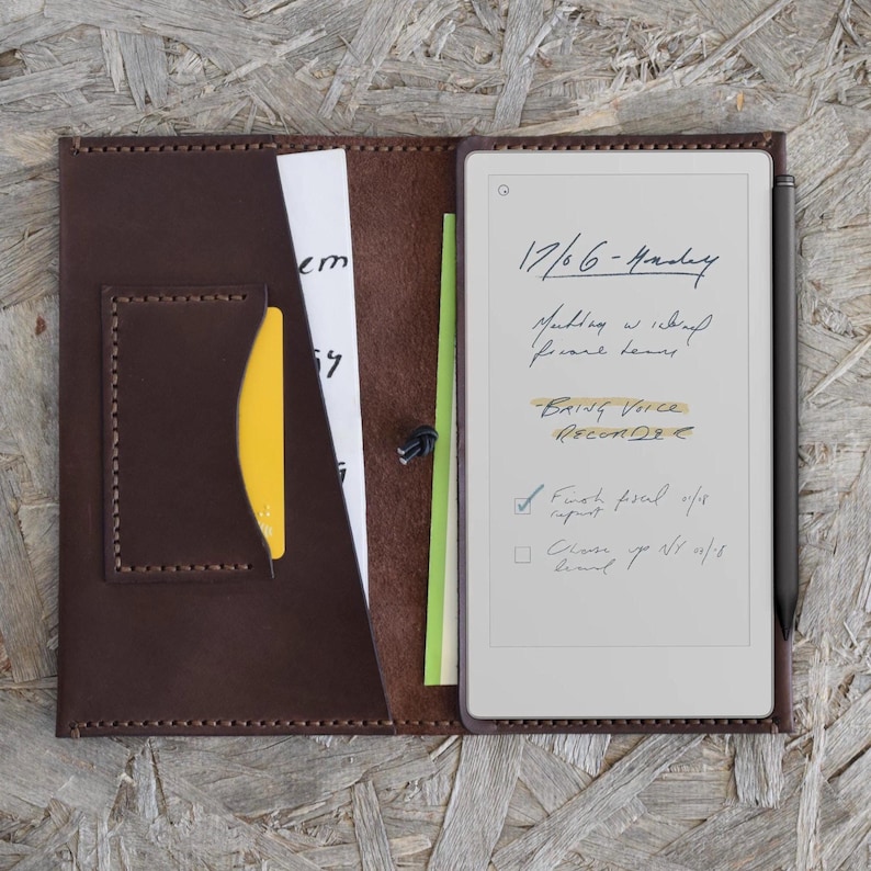 May include: A brown leather folio with a digital notepad and stylus. The notepad displays handwritten notes, including the date, "17/6 - Monday", and a to-do list. A yellow card and other papers are visible in the folio.