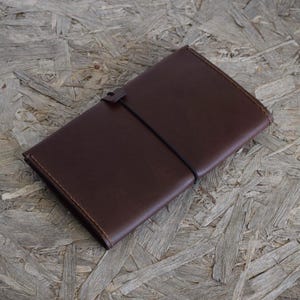 May include: A dark brown leather journal with a simple, elegant design. The cover is smooth and rectangular, with a strap closure. The journal is closed with a black elastic band. The edges are neatly stitched, and the overall aesthetic is minimalist and classic.
