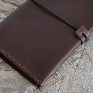 May include: A dark brown leather journal with a stitched border and a black elastic closure. The journal is rectangular and appears to be closed. The background is a light brown wood surface.