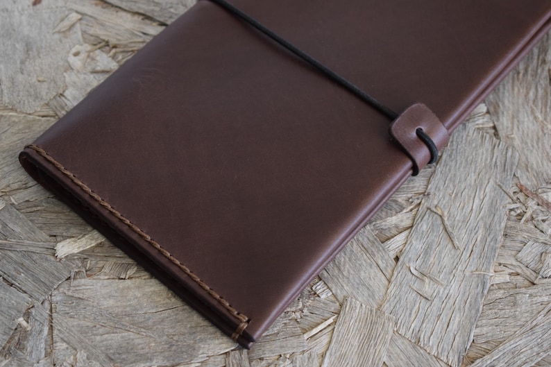 May include: A dark brown leather journal with a stitched border and a black elastic closure. The journal is rectangular and appears to be closed. The background is a light brown wood surface.