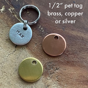 May include: 1/2 inch pet tag in brass, copper, or silver. The tag is round and has a hole for a ring. The tag is shown on a wooden surface. The tag is blank except for the word "MAX" on one of the tags.