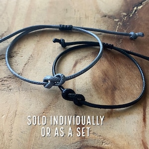 May include: Two friendship bracelets, one gray and one black, tied in a knot. The bracelets are on a wooden surface. "Sold individually or as a set" is written below the bracelets.