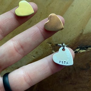 Pu&ograve; includere: Tre etichette di metallo a forma di cuore. Due sono lisce, una dorata e una in rame. La terza etichetta &egrave; in argento e ha il nome "PITA" inciso su di essa.