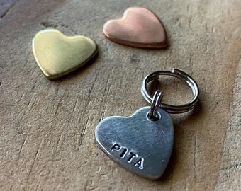 May include: A silver heart-shaped dog tag with the name "PITA" engraved on it. The tag is attached to a silver key ring. The tag is sitting on a wooden surface next to two other heart-shaped tags, one gold and one copper.