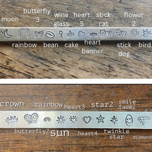 May include: Close-up of two metal stamp sets with various symbols and words. The top set includes symbols for a moon, rainbow, butterfly, coffee bean, wine glass, cake, heart, cat, stick, flower, and bird. The bottom set includes symbols for a crown, butterfly, rainbow, sun, heart, star, and diamond. The words on the sets include "moon", "butterfly", "wine", "heart", "stick", "flower", "rainbow", "bean", "cake", "heart", "banner", "stick", "bird", "dog", "crown", "rainbow", "heart", "star", "smile", "face", "butterfly", "sun", "heart", "twinkle", "star", and "diamond".