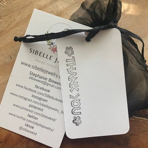Pu&ograve; includere: Un biglietto bianco con testo nero che dice "SIBELLE JEWELRY" e "MAKING THINGS PRETTY". Il biglietto include anche un indirizzo del sito web e le maniglie dei social media per l'azienda. Il biglietto &egrave; legato a una borsa di organza nera con un timbro bianco "THANK YOU".