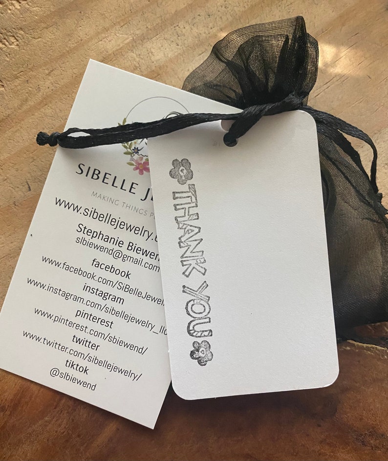 Pu&ograve; includere: Un biglietto bianco con testo nero che dice "SIBELLE JEWELRY" e "MAKING THINGS PRETTY". Il biglietto include anche un indirizzo del sito web e le maniglie dei social media per l'azienda. Il biglietto &egrave; legato a una borsa di organza nera con un timbro bianco "THANK YOU".