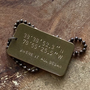 May include: A brass dog tag with a chain. The tag has the coordinates "38.38, 52.3... N 76.55, 25.5... W" and the text "Where it all began".