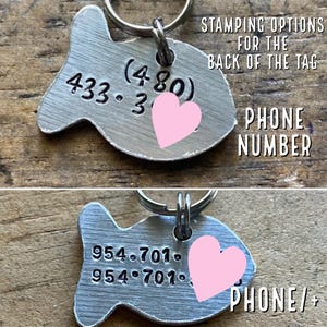 May include: Two silver fish-shaped pet ID tags with key rings. The top tag has the phone number 433-3 (480) and a pink heart. The bottom tag has the phone number 954-701 and a pink heart. The text "STAMPING OPTIONS FOR THE BACK OF THE TAG" is above the top tag. The text "PHONE NUMBER" is to the right of the top tag. The text "PHONE/+" is to the right of the bottom tag.